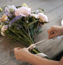 Händer knyter ett band runt en bukett med rosor och andra blommor på ett träbord.